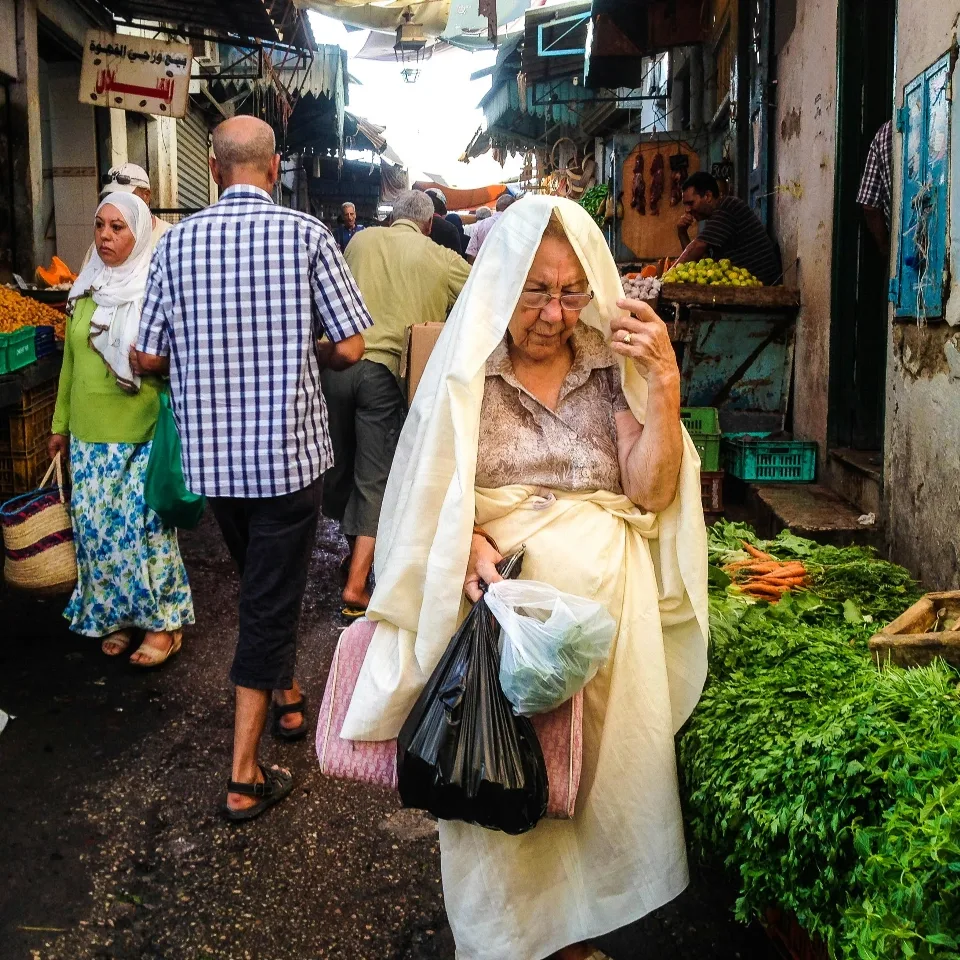 Shoppers at a Tunisian market, a woman in a traditional sefseri walking past vegetable stalls